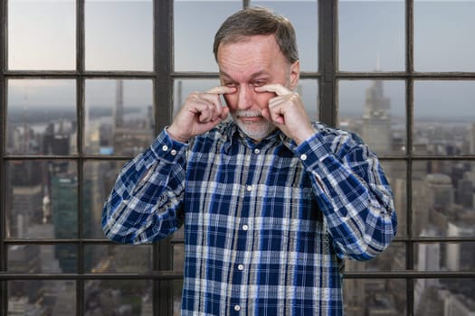 Portrait of an aged man rubbing his eyes. Tired of office work. Checkered window background with urban scape view.