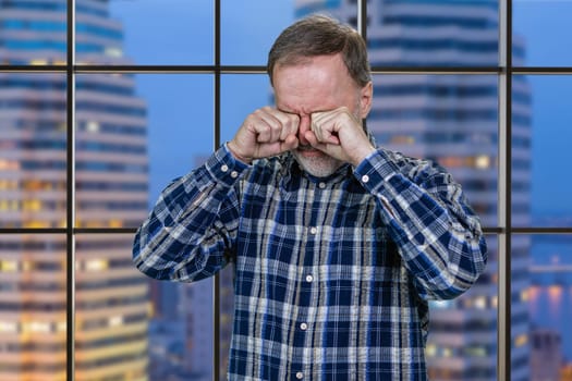 Portrait of crying aged senior man is wiping his tears. Checkered windows with night cityscape background.