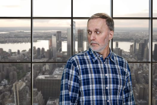 Portrait of confused aged man is looking back. Checkered window background with cityscape view.