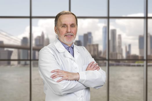 Portrait of cheerful senior doctor with folded arms. Checkered window background with cityscape view.