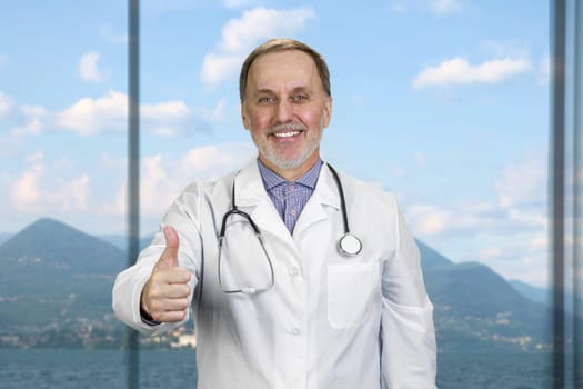 Happy smiling senior doctor with stethoscope shows thumb up. Window with landscape view in the background.