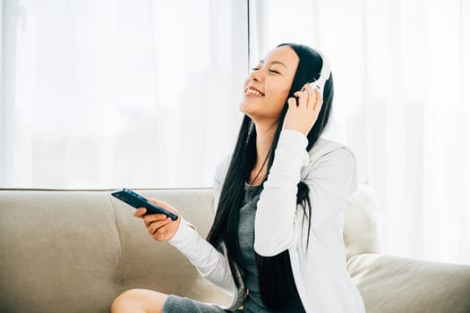 Young woman on sofa enjoys leisure time listens to music using smartphone and headphones. Embracing relaxation enjoyment and modern technology at home.