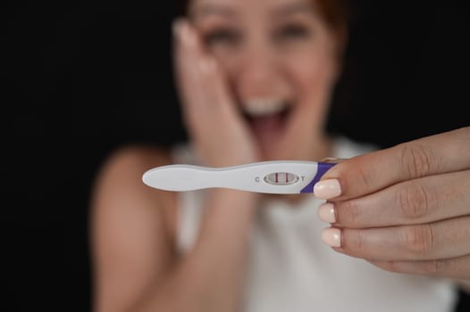 Blurred portrait of happy caucasian woman holding positive express pregnancy test on black background