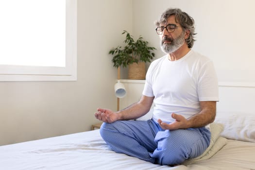 Mature adult man relaxing, doing meditation sitting on bed at home. Male meditating in the morning. Copy space. Wellness and mental health concept.