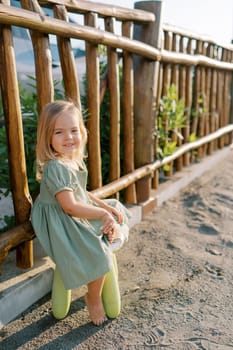 Little smiling girl sitting on a plastic stool near a wooden fence on a playground. High quality photo