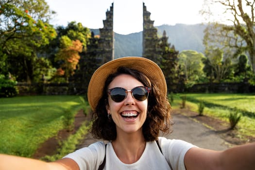 Happy young woman traveler looking at camera taking selfie in Balinese Hindu Temple entrance during summer vacation. Indonesia. Travel, freedom and happiness concept.