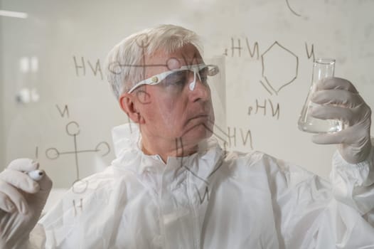 An elderly Caucasian male chemist in a protective suit looks at a test tube and writes formulas on a glass wall
