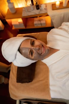 Serene ambiance of spa salon, top view woman customer indulges in rejuvenating with charcoal face cream massage with warm lighting candle. Facial skin treatment and beauty care concept. Quiescent