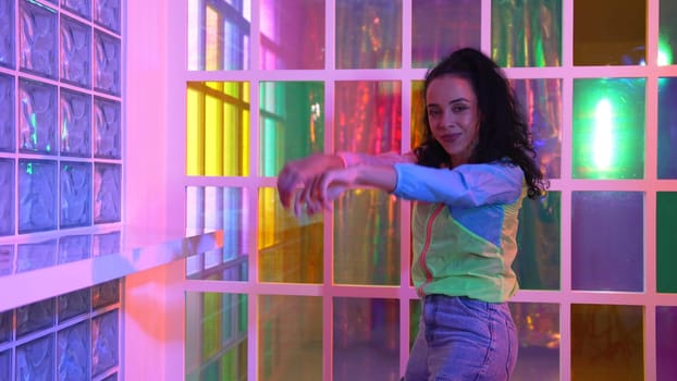 Happy hispanic girl in lively mood moving along music in neon light while wearing colorful cloth at night club. Street dancer making energetic footstep or movement while looking at camera. Regalement.