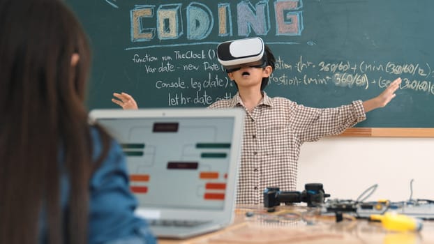 Asian boy wearing VR headset to enter metaworld at table with electronic board while girl coding system. Group of diverse student working together to develop program and writing engineering code. Pedagogy.