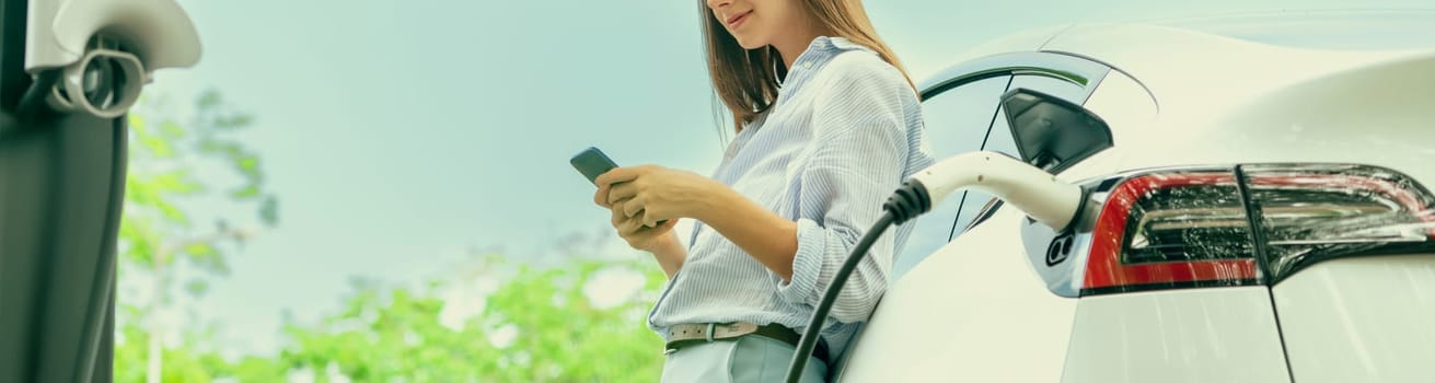 Young woman using smartphone online banking application to pay for electric car battery charging from EV charging station during vacation road trip at national park or summer forest. Panorama Exalt