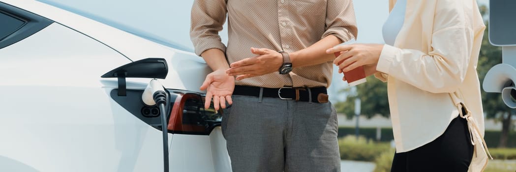 Young couple with coffee recharge EV car battery at charging station connect to electrical industrial power grid. Couple with shopping and travel using eco electric car lifestyle.Panorama Expedient