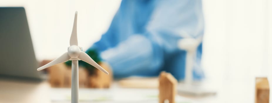 Windmill model represented using clean energy and wooden block represented eco house was scatter around on the table in front of businessman working on laptop. Blurring background. Delineation.