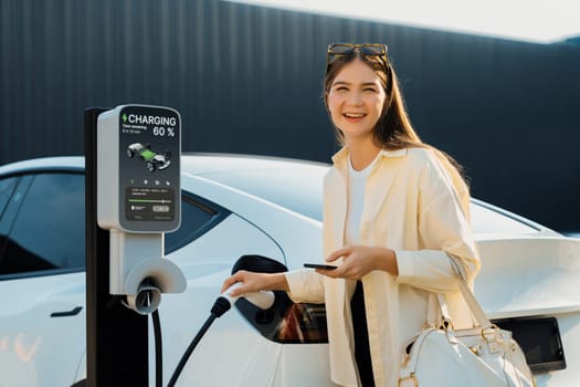 Young woman holding shopping bag and use smartphone to pay for electricity for recharging EV car battery from charging station at city mall parking lot. Modern woman go shopping by eco car. Expedient