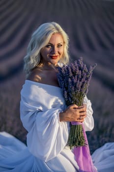 Blonde woman poses in lavender field at sunset. Happy woman in white dress holds lavender bouquet. Aromatherapy concept, lavender oil, photo session in lavender.