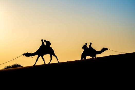 Silhouette of camel with two people sitting on it crossing over sand dunes in Sam Jaisalmer Rajasthan India