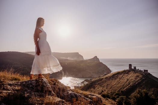 A woman stands on a rocky hill overlooking the ocean. She is wearing a white dress and she is enjoying the view. The scene is serene and peaceful, with the sun shining brightly in the background