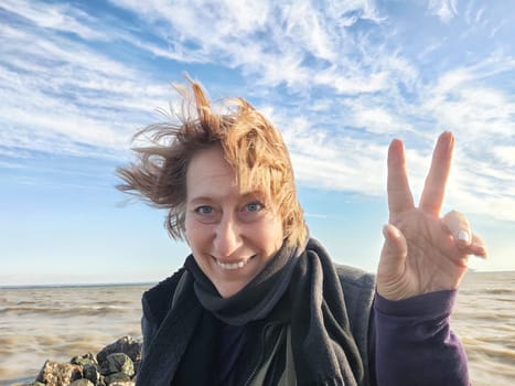 Happy cheerful middle aged woman in scarf taking selfie on nature outdoors and sun with water and waves of sea on the background