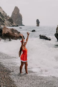 Woman travel sea. Young Happy woman in a long red dress posing on a beach near the sea on background of volcanic rocks, like in Iceland, sharing travel adventure journey