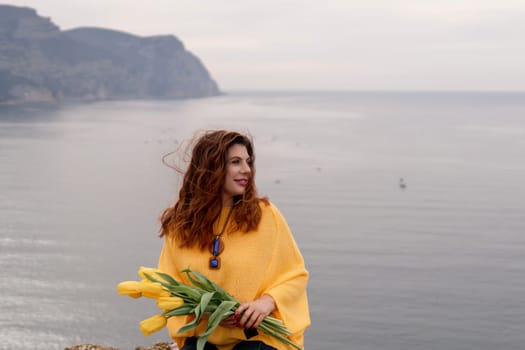 Portrait happy woman woman with long hair against a background of mountains and sea. Holding a bouquet of yellow tulips in her hands, wearing a yellow sweater.