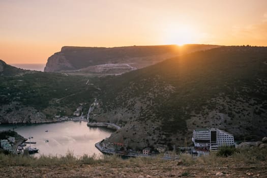 A beautiful sunset over a mountain with a small town in the distance. The sun is setting behind the mountains, casting a warm glow over the landscape. The water in the valley is calm and peaceful