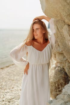 woman white dress standing on a beach, looking out at the ocean. She is lost in thought, with her hand on her hip. Concept of calm and introspection