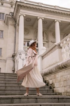 A woman in a hat stands on a set of stairs in front of a building. She is wearing a coat and a skirt