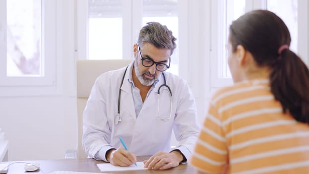 Mature doctor talking with a woman while writing down notes sitting on a desk in a clinic