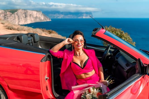 A woman is sitting in the back of a red convertible car. The car is parked on a dirt road near the ocean. The woman is wearing a pink suit and red shoes. Scene is relaxed and carefree