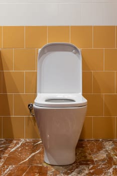 Opened lid of rectangular toilet seat made of white plastic on marble flooring in a bathroom with yellow tiled walls.