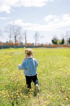 Little girl in a wreath of dandelions walks along a flowering lawn. Back view. High quality photo