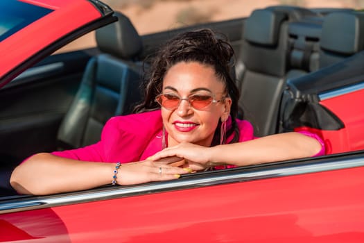 A woman in a pink jacket is sitting in a red convertible. She is wearing sunglasses and has her hand on her hip. Scene is relaxed and carefree