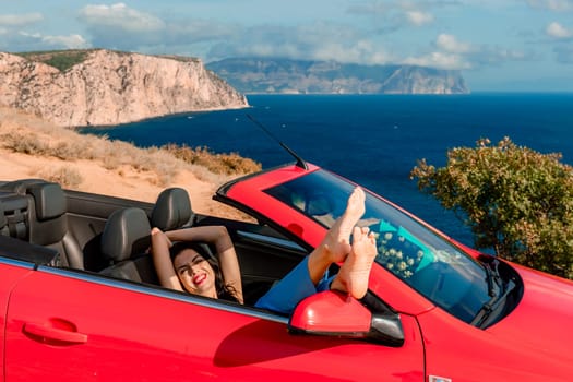 A woman is laying in the back of a red convertible car, with her feet up in the air. The car is parked on a cliff overlooking the ocean. Concept of freedom and relaxation