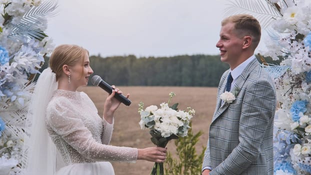 The bride and groom take their vows with a microphone in front of the wedding arch