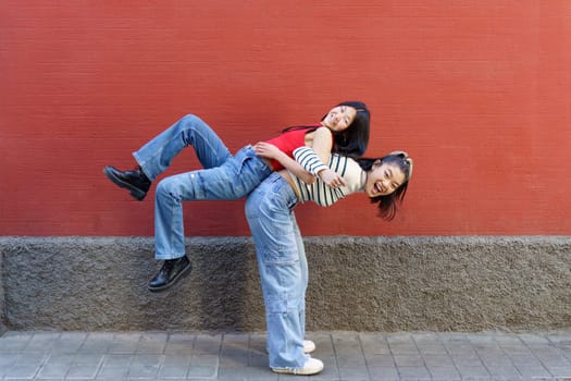 Side view of cheerful Asian girl in casual clothes lifting smiling friend on back while standing against red wall
