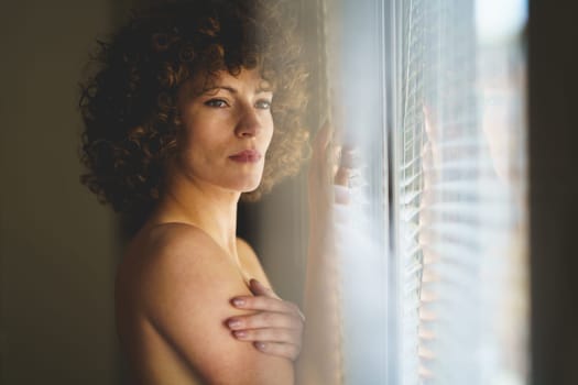 Side view of pensive young nude woman with curly hair, leaning on window and looking away thoughtfully.