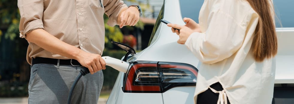 Young couple use smartphone to pay for electricity at public EV car charging station green city park. Modern environmental and sustainable urban lifestyle with EV vehicle. Panorama Expedient