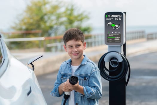 Playful little boy pointing EV charger at camera, recharging eco-friendly electric car from EV charging station. EV car travel by the seashore using clean and sustainable energy.Perpetual