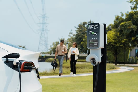 Young couple with coffee recharge EV car battery at charging station connected to electrical industrial power grid tower. Couple with shopping and travel using eco electric car lifestyle. Expedient