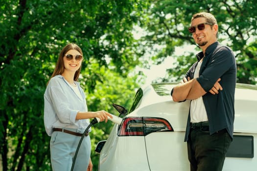 Lovely young couple wearing sun glasses recharging battery for electric car during road trip travel EV car in natural forest or national park. Eco friendly travel during vacation and holiday. Exalt