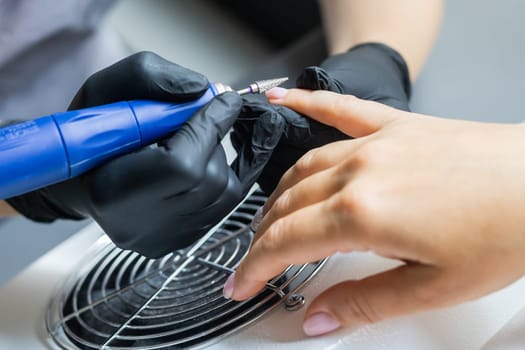 Female master using electric nail file while doing manicure for client in beauty salon.