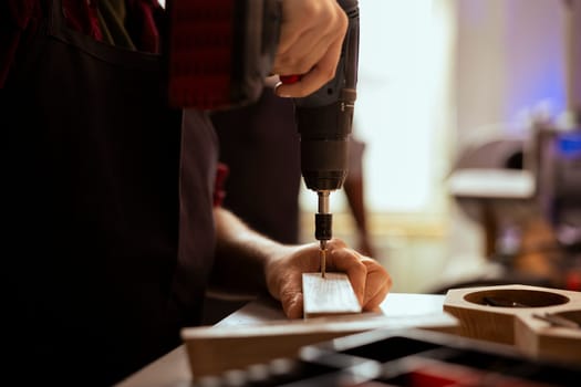 Carpenter drilling hole into timber block, doing furniture assembling in studio, close up. Woodworking expert in joinery inserting nail into piece of wood using electric power drill tool