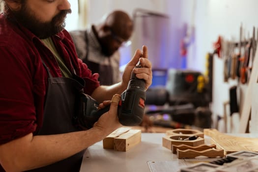 Carpenter changing power drill head into suitable one for task ahead, making holes into timber block. Cabinetmaker in assembly shop preparing electric gear tool to use for commissioned job