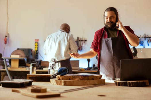 Smiling manufacturer discussing on telephone call with client inquiring about furniture assembling commission progress. Cheerful carpenter in assembly shop talking on mobile phone with customer