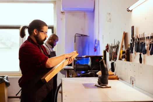 Carpenter wearing protection glasses, checking for scratches on wood before assembling furniture. Cabinetmaker using safety equipment in joinery, running hand along lumber block, running evaluation