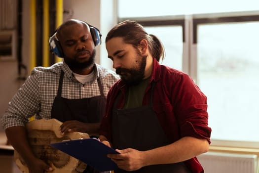 Carpenter and african american colleague verifying piece of wood before using it for furniture assembly. Cabinetmaker and apprentice inspecting lumber block for damages, crosschecking with checklist