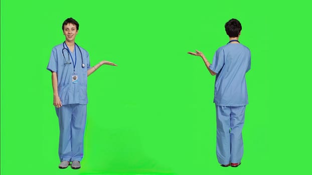 Medical assistant pointing at something aside and doing web commercial, creating advertisement against greenscreen backdrop. Young nurse with scrubs showing a thing to left or right sides. Camera A.