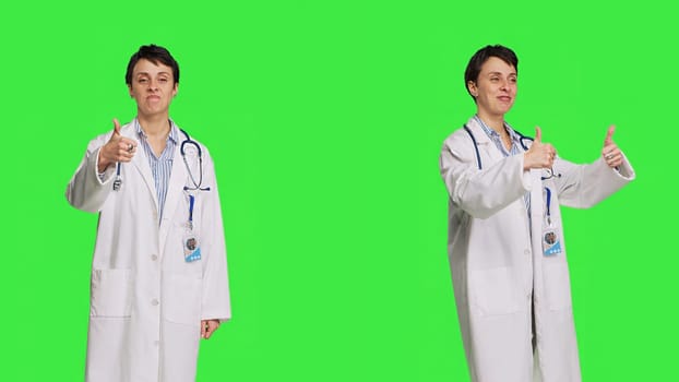 Cheerful medic doing thumbs up symbol against greenscreen backdrop, expresses positivity with like sign. General practitioner with coat giving approval and being satisfied with success. Camera B.