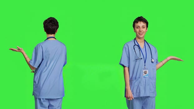 Medical assistant pointing at something aside and doing web commercial, creating advertisement against greenscreen backdrop. Young nurse with scrubs showing a thing to left or right sides. Camera B.