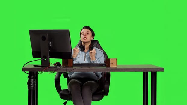 Young adult playing video games on computer at desk against greenscreen backdrop, winning worldwide tournament on pc. Woman gamer feeling pleased and happy with her success. Camera B.